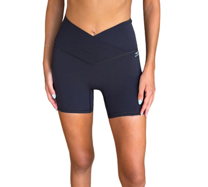 A model wearing high-waisted blackathletic biker shorts with a V-cut waist, paired with a white sports bra, standing in a modern indoor setting with a wicker sofa in the background.