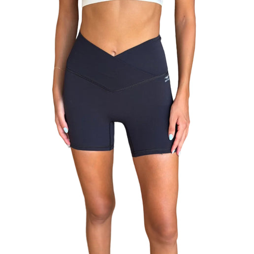 A model wearing high-waisted blackathletic biker shorts with a V-cut waist, paired with a white sports bra, standing in a modern indoor setting with a wicker sofa in the background.